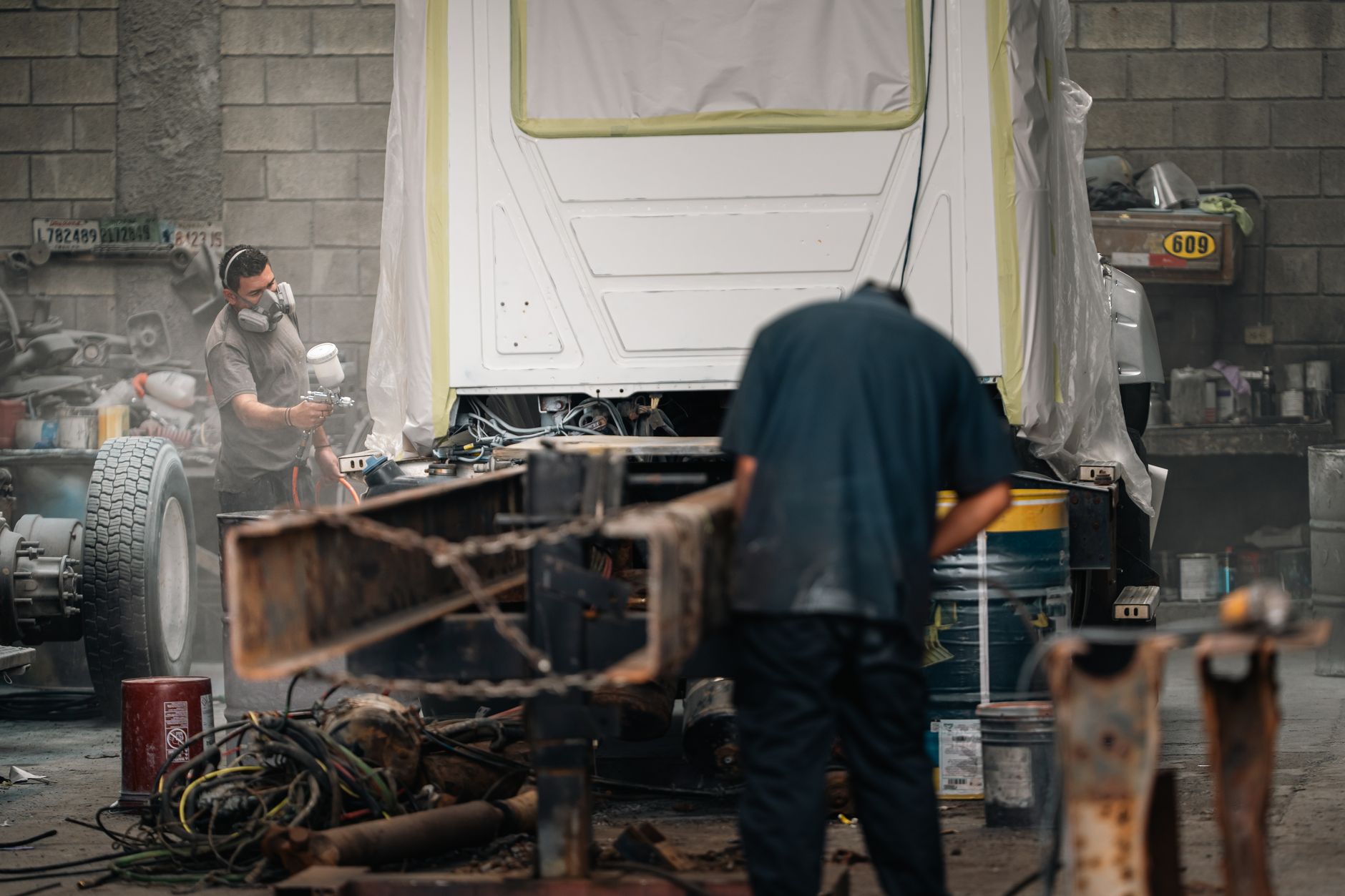 Two workers in protective gear painting a vehicle chassis in a body shop